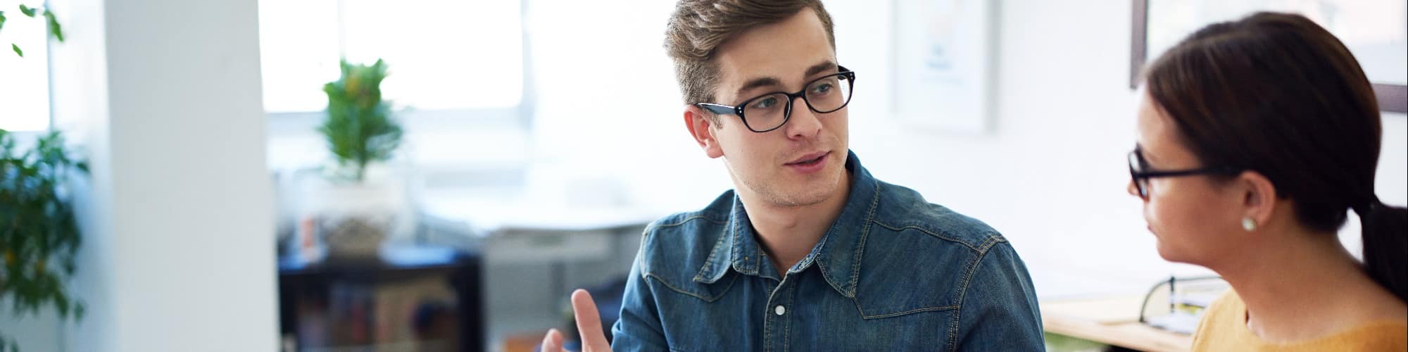 a person wearing glasses and a denim shirt discussing with a woman